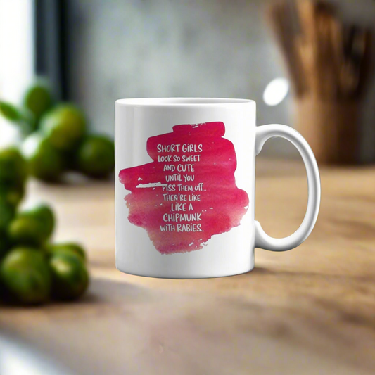 The Short Girls Ceramic Mug by Kreative Kreationz features a pink watercolor splash and the quote, "Short girls look so sweet and cute until you piss them off. They’re like a chipmunk with rabies." Dishwasher safe for easy cleaning.