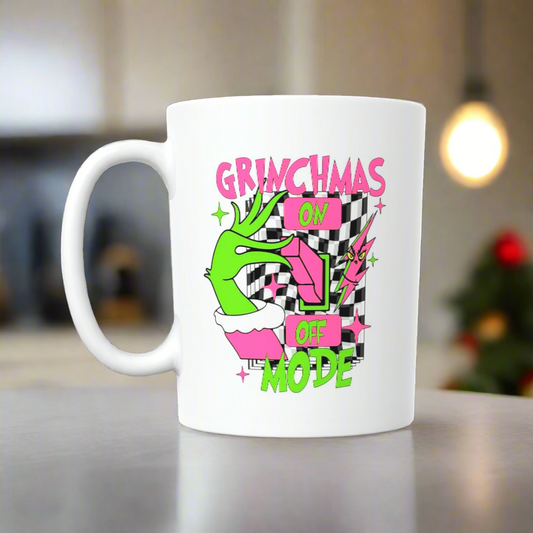 The Grinchmas Ceramic Mug by Kreative Kreationz features a green hand flipping a switch with “GRINCHMAS ON OFF MODE” in pink and green on a black-and-white checkered background—perfect for celebrating Grinchmas mode!.