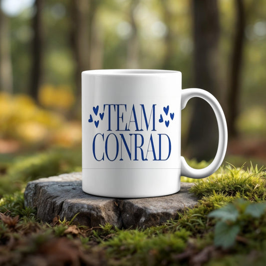A "TEAM CONRAD" white mug with blue hearts rests on a tree stump in the woods—pair it with the Kreative Kreationz (Copy) Team Conrad Inspired Keychain for the perfect gift for fans.