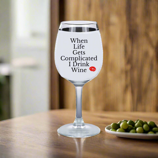 A wine glass with white liquid rests on wood, featuring "When Life Gets Complicated I Drink Wine" and a red lips design. Nearby is a dish of green olives and a Kreative Kreationz neoprene Wine Koozie, perfect for any wine enthusiast.
