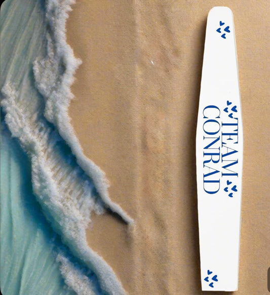 A white surfboard with blue paw prints and "TEAM CONRAD" rests on the sandy beach by foamy waves, next to the Kreative Kreationz Team Conrad Ceramic Nail File—an eco-friendly, washable, and reusable nail file.
