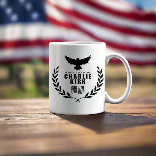 The Kreative Kreationz Charlie Kirk Forever In Our Hearts Ceramic Mug rests on wood, displaying an eagle, laurel wreath, small American flag, and heartfelt text, with a blurred flag behind—a meaningful tribute keepsake.