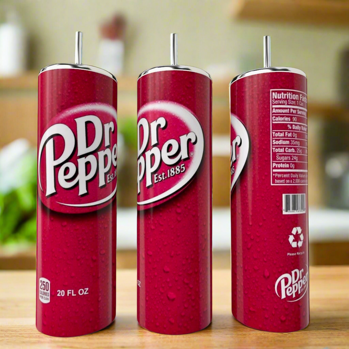 A Dr Pepper Stainless Steel Tumbler by Kreative Kreationz sits on a kitchen counter with three tall Dr Pepper cans nearby, showing the logo, nutrition facts, and recycling symbol against a blurred kitchen background.