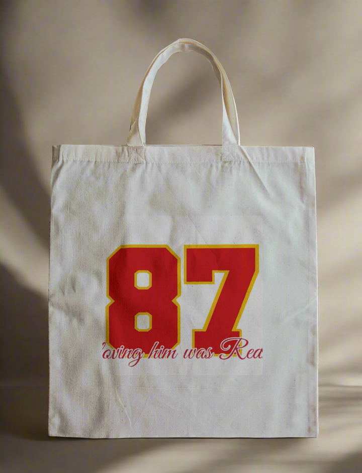 Show your spirit with the "Loving Him Was Red" reusable canvas bag by Kreative Kreationz, featuring bold red and yellow "87" graphics and cursive text—a stylish way to carry your essentials.