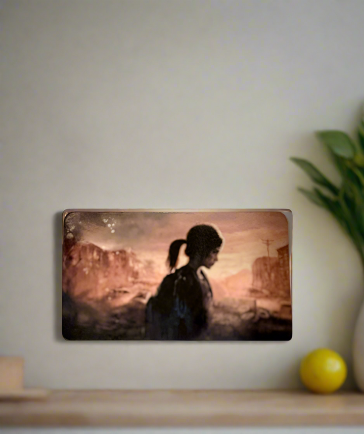 A portrait of a ponytailed figure in profile hangs on a beige wall. Nearby, gaming collectibles stand out—a Kreative Kreationz The Last Of Us Fridge Magnet sits on the shelf beside a green plant and yellow ball against a desolate landscape backdrop.