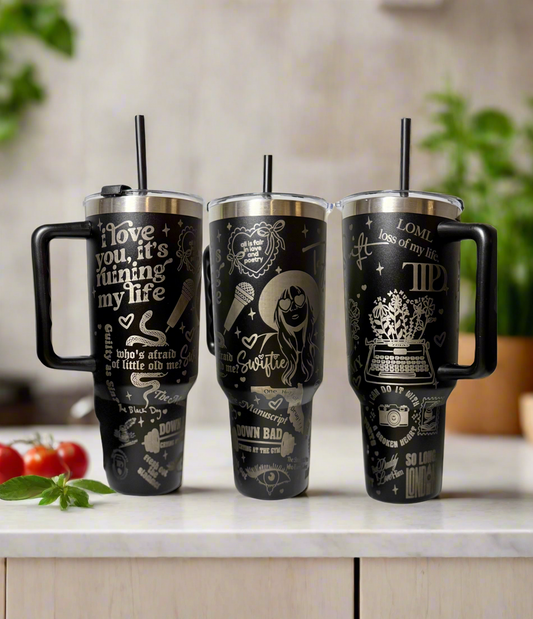 Three large black Kreative Kreationz Taylor Swift TTPD 40oz Engraved Tumblers with handles sit on a kitchen counter. Each stainless steel mug has white Taylor Swift-themed designs. A blurred kitchen scene with tomatoes is in the background.