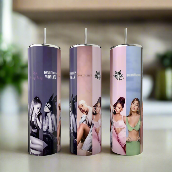 Three Kreative Kreationz Ariana Grande All Albums 20oz Tumblers, featuring images of Ariana in various poses and outfits, are placed on a table against a blurred indoor background.