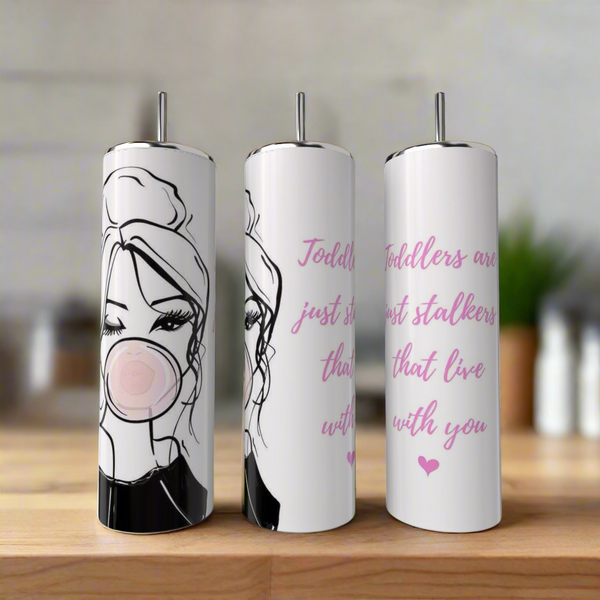 Three 20oz tumblers by Kreative Kreationz sit on wood: the left shows a woman with a bun and bubblegum; the other two luxury tumblers feature pink hearts and the phrase, “Toddlers are just short stalkers that live with you.”.