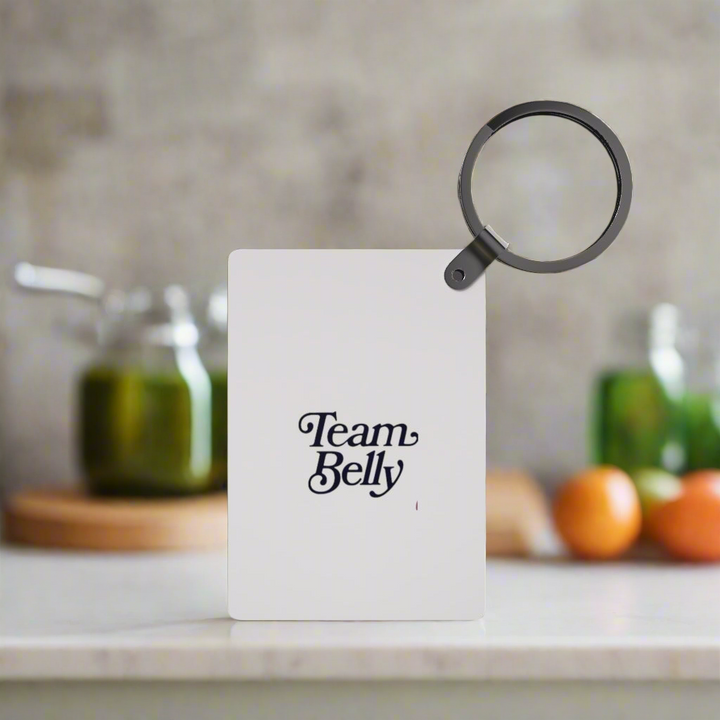A Team Belle Inspired Keychain from Kreative Kreationz is displayed upright on a kitchen counter, making a perfect gift. Jars and vegetables are blurred in the background for a cozy touch.