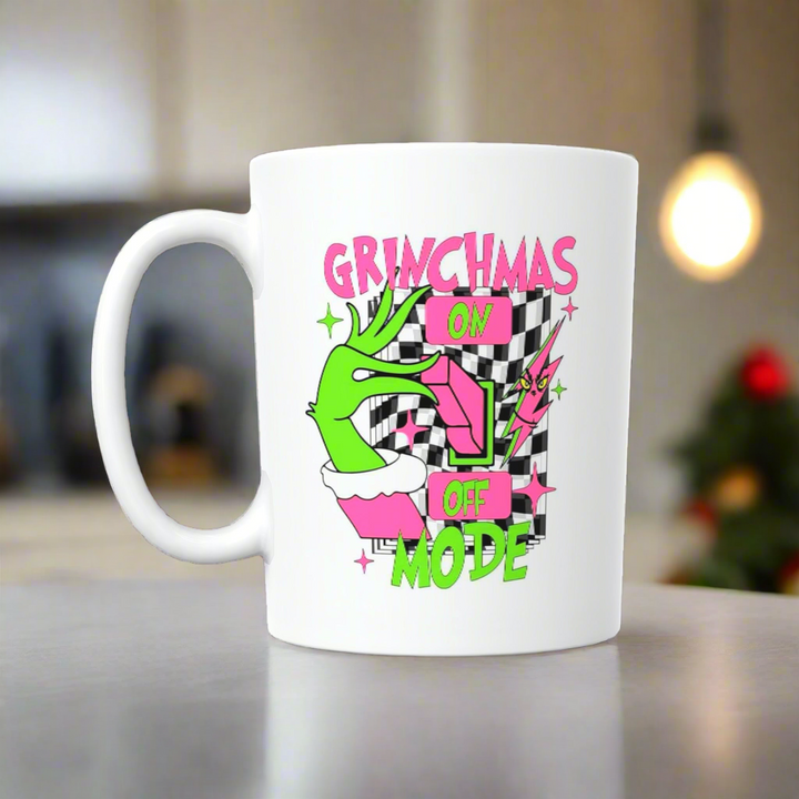 The Grinchmas Ceramic Mug by Kreative Kreationz features a green hand flipping a switch with “GRINCHMAS ON OFF MODE” in pink and green on a black-and-white checkered background—perfect for celebrating Grinchmas mode!.