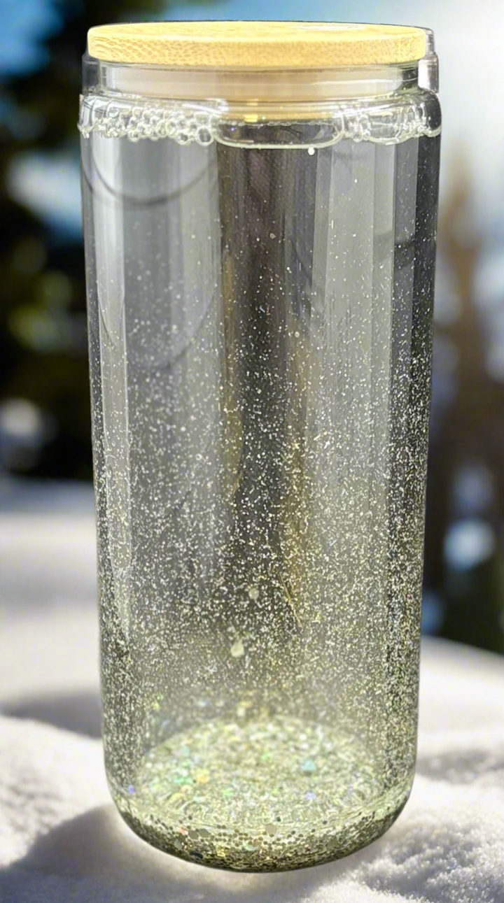 The Snowglobe Tumbler by Kreative Kreationz, a clear glass glitter tumbler with bamboo lid, sits on a white surface as sunlight shines through, making the glitter sparkle and float in the water.
