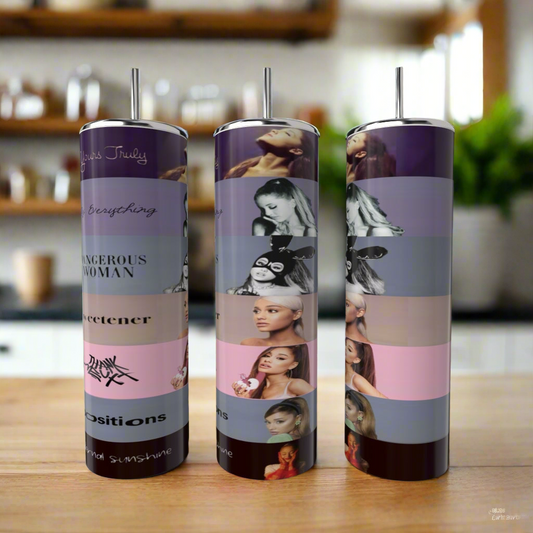 Three Kreative Kreationz Ariana Grande 20oz tumblers with metal straws sit on a kitchen counter, each decorated in vibrant horizontal bands featuring artistic portraits, song titles, and album covers.
