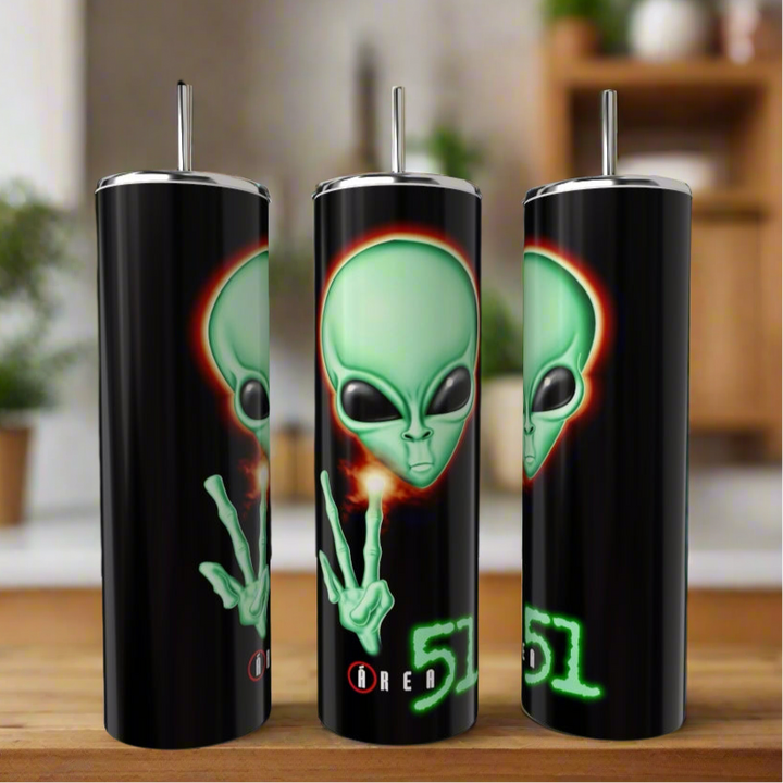 Three black Area 51 20oz Skinny Tumblers by Kreative Kreationz feature a large green alien face making a peace sign, glowing “Area 51” text, and come with reusable metal straws. The eco-friendly tumblers are shown indoors on a wooden surface.
