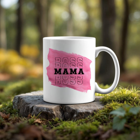The Mama Mug by Kreative Kreationz features "BOSS MAMA" over a pink brushstroke. Shown on a tree stump amid forest moss, this ceramic mug is dishwasher safe and blends stylish design with natural scenery.