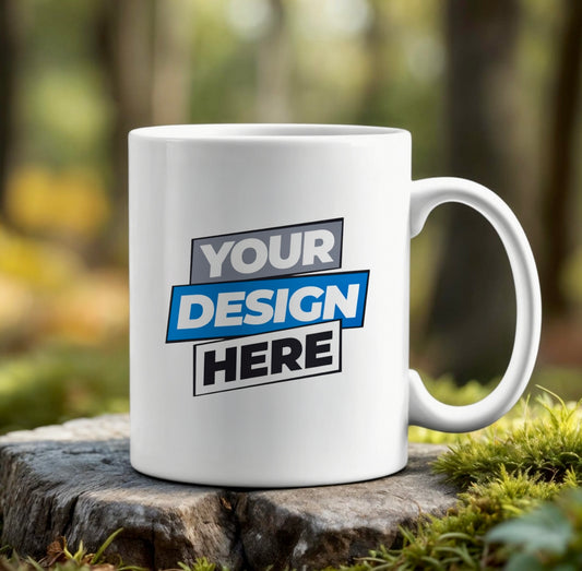 The Kreative Kreationz Custom Ceramic Mug (15oz | 11oz) features "Your Design Here" in bold black, gray, and blue text. Shown outdoors on a tree stump with a blurred forest background. Dishwasher safe—upload your own design!.
