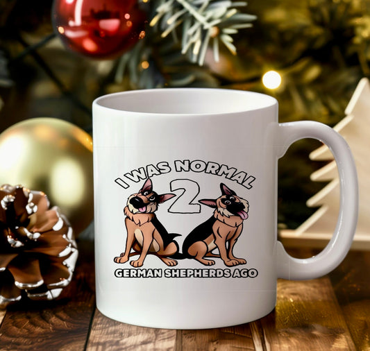 A white ceramic mug from Kreative Kreationz sits on a wooden table amid festive Christmas decor. Featuring two cartoon German Shepherds and the text, "I WAS NORMAL 2 GERMAN SHEPHERDS AGO," this German Shepherd Ceramic Mug captures a playful custom spirit.