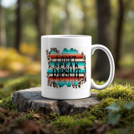 The Yellowstone Ceramic Mug by Kreative Kreationz, featuring the bold text "I DON'T SPEAK DIPSHIT," rests on a mossy tree stump outdoors amid vibrant greenery and blurred foliage in the background.