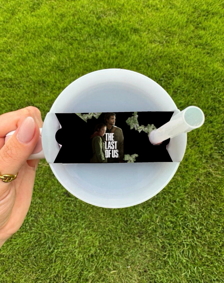 A hand holds a white cup with a straw and the Kreative Kreationz The Last Of Us Stanley Lid Topper, featuring "The Last of Us" characters and logo on top. The background is green grass.