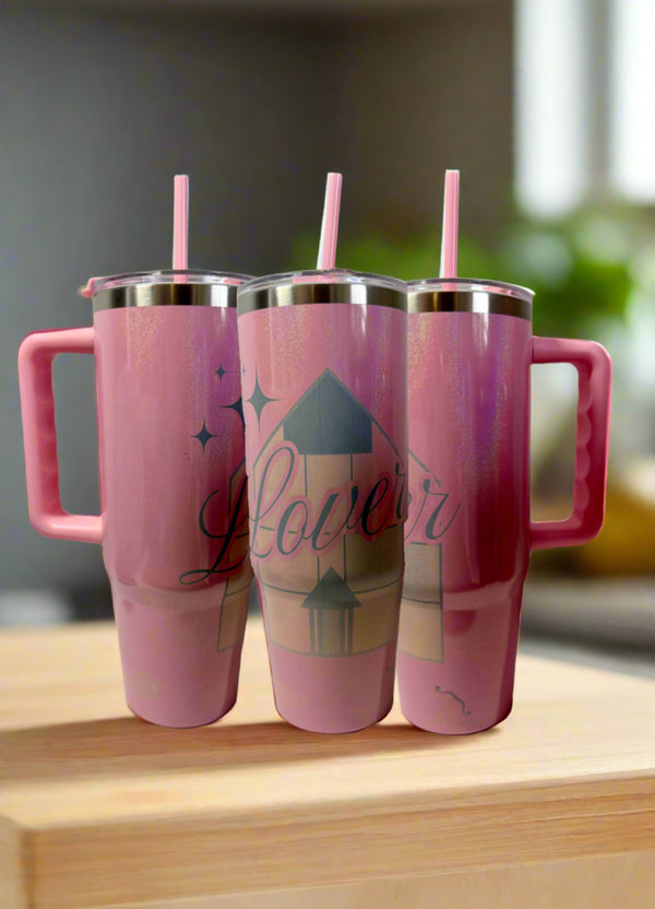 Three pink Kreative Kreationz stainless steel tumblers with handles and straws are displayed on wood. The center "Lover House Taylor Swift Engraved Tumbler" features a house design and "Lover" in cursive, set against a softly blurred background.