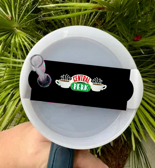 A close-up of the Friends Stanley Lid Topper by Kreative Kreationz features a white circular lid with the "Central Perk" logo and steaming coffee cups. A clear tube extends from it, set against lush greenery, highlighting craftsmanship with premium materials.