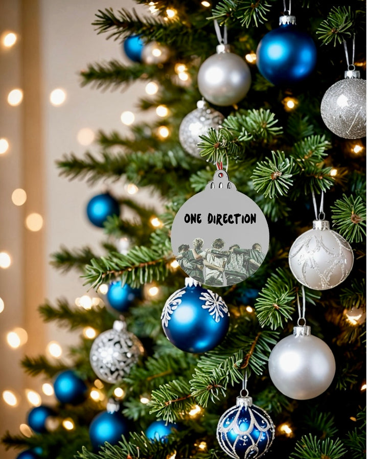 A Christmas tree decorated in blue, silver, and white features the Kreative Kreationz One Direction Christmas Tree Ornament, displaying "ONE DIRECTION" and five illustrated figures amid glowing string lights.