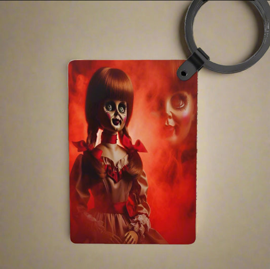 The Annabelle Inspired Keychain by Kreative Kreationz features a creepy porcelain doll with wide dark eyes and braided hair, dressed in red and white against a smoky red background—a unique, durable personalized gift.