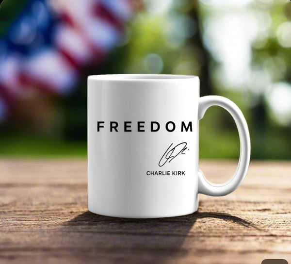The Freedom Charlie Kirk Mug by Kreative Kreationz sits on wood, featuring "FREEDOM," a signature, and "CHARLIE KIRK." Bright natural light and a blurred American flag complete this ideal patriotic tribute gift.
