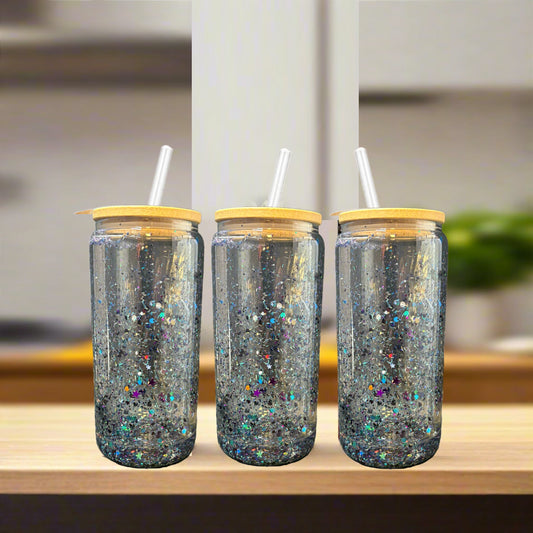 Three Kreative Kreationz Snowglobe Tumblers—16oz clear glass with bamboo lids and straws—are filled with sparkling glitter and sit side by side on a light wooden surface, against a blurred kitchen background.