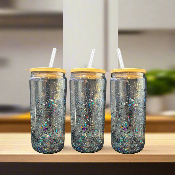 Three Kreative Kreationz Snowglobe Tumblers—16oz clear glass with bamboo lids and straws—are filled with sparkling glitter and sit side by side on a light wooden surface, against a blurred kitchen background.